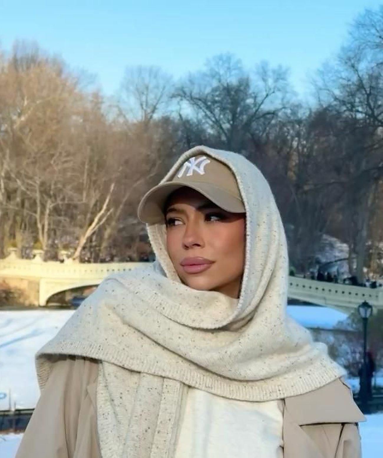 Kayla Richart in a cream knit hood and tan cap, snow and a stone bridge in Central Park behind her